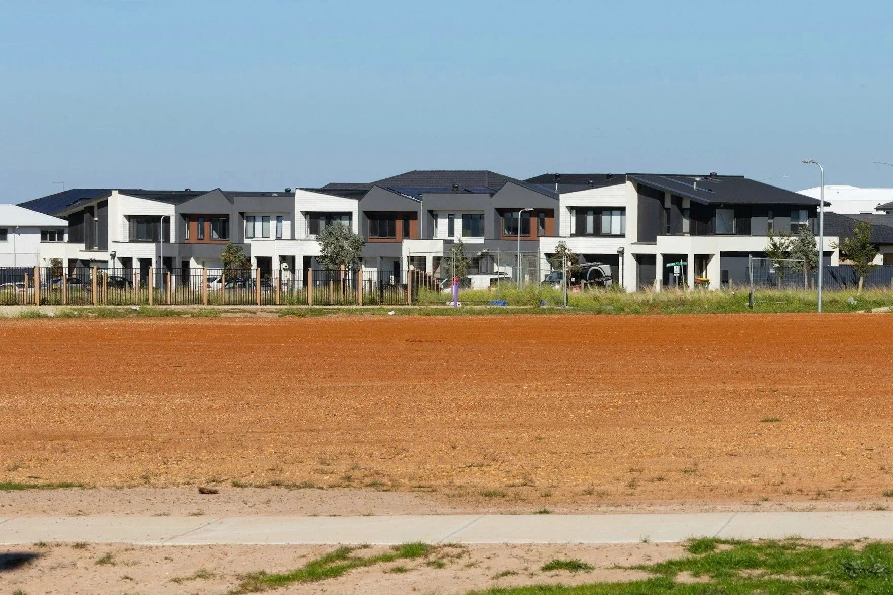 Modern townhouses with green trees, parked cars, and street lamps, viewed behind a fence from a dirt and grassy area