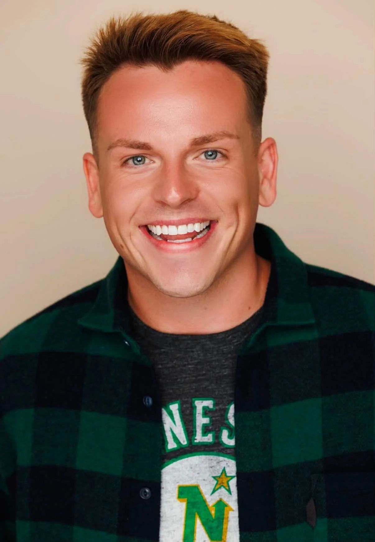 A young man with short brown hair and blue eyes, smiling with white teeth, wearing a dark green and black plaid shirt layered over a gray t-shirt with green, white, and yellow print, standing against a neutral background.