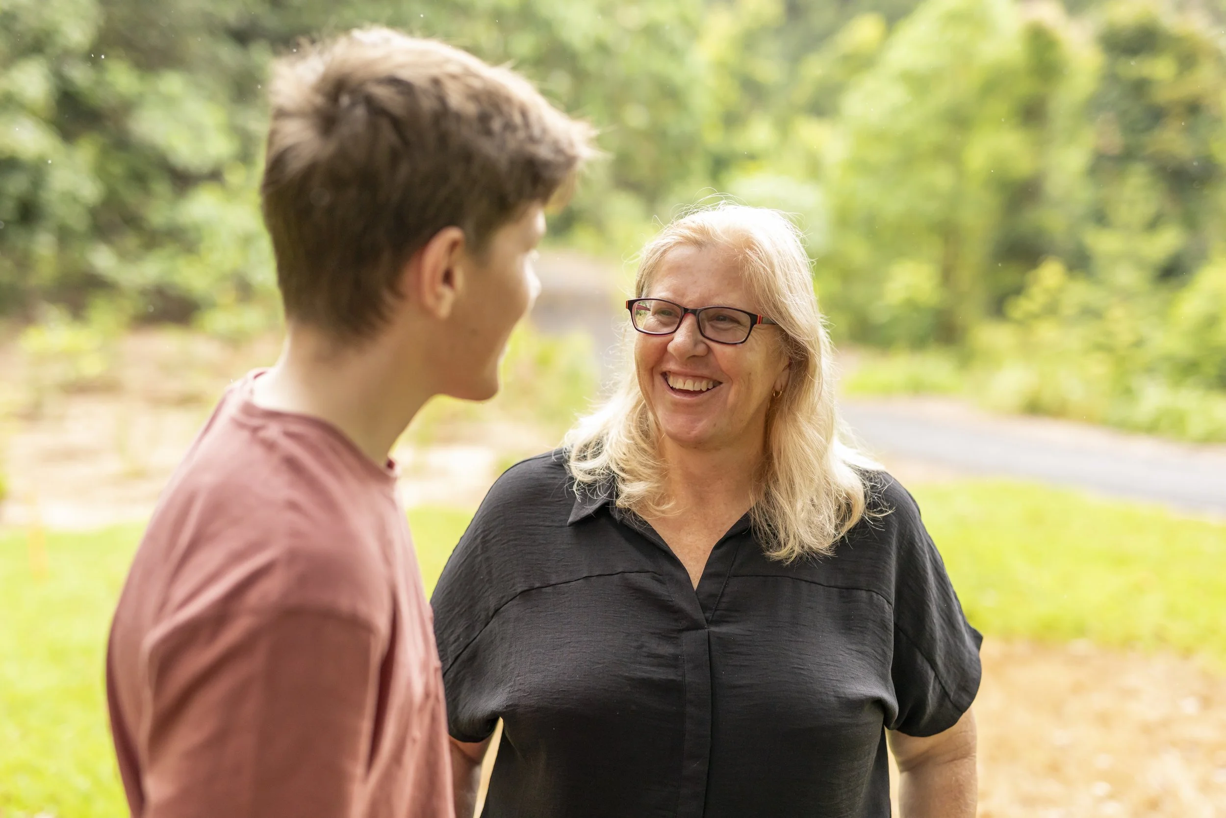 A woman with blond hair and glasses smiling and talking to a young man with short brown hair, outdoors on a sunny day with green trees in the background.