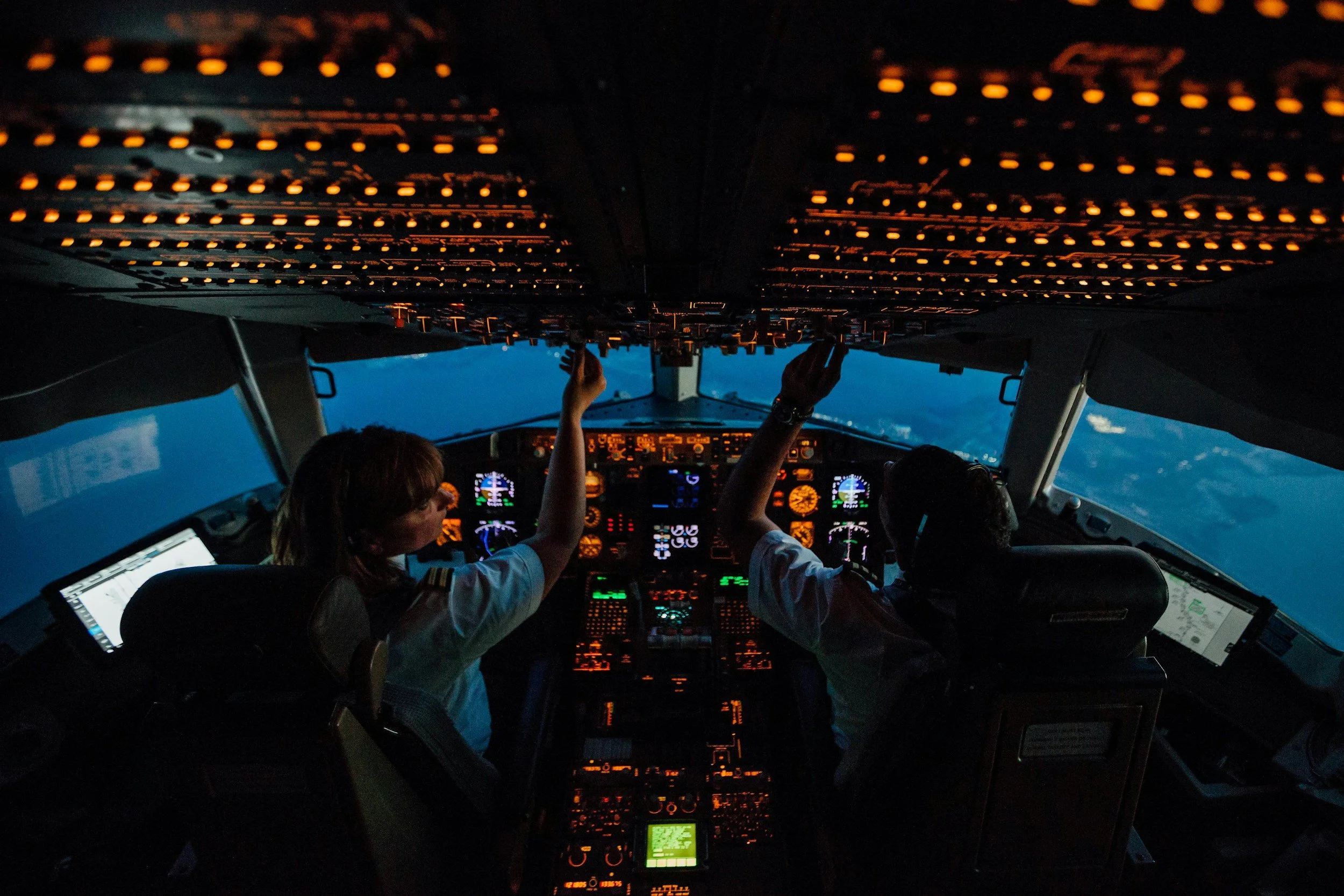 Inside the cockpit of an airplane during flight, showing pilots operating the controls with a view of the sky outside the windshield at dusk or dawn.