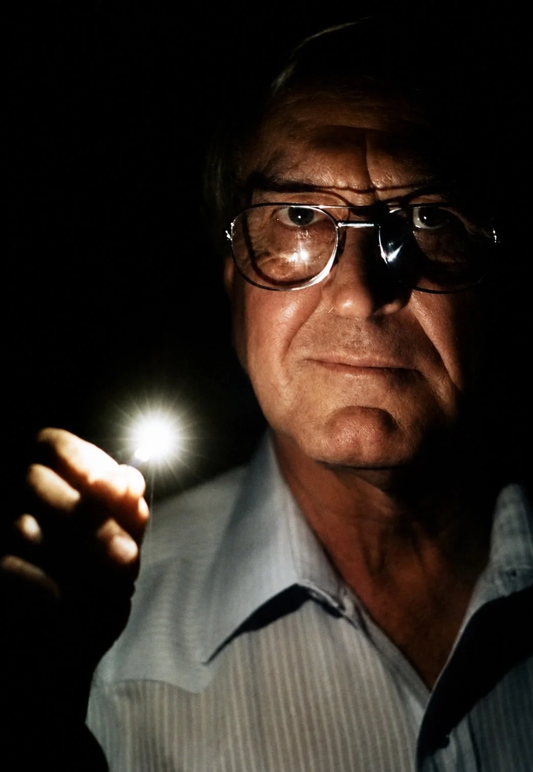 A man wearing glasses holding a small flashlight in a dark setting, with the light illuminating part of his face.