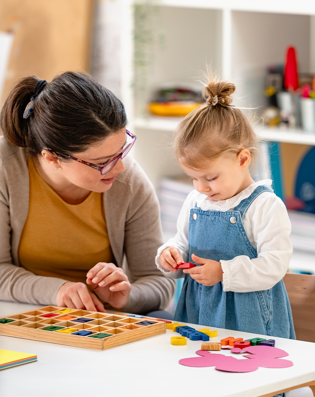 A young girl with a top knot hairstyle playing a colorful educational game with an adult woman at a table in a classroom or play area.