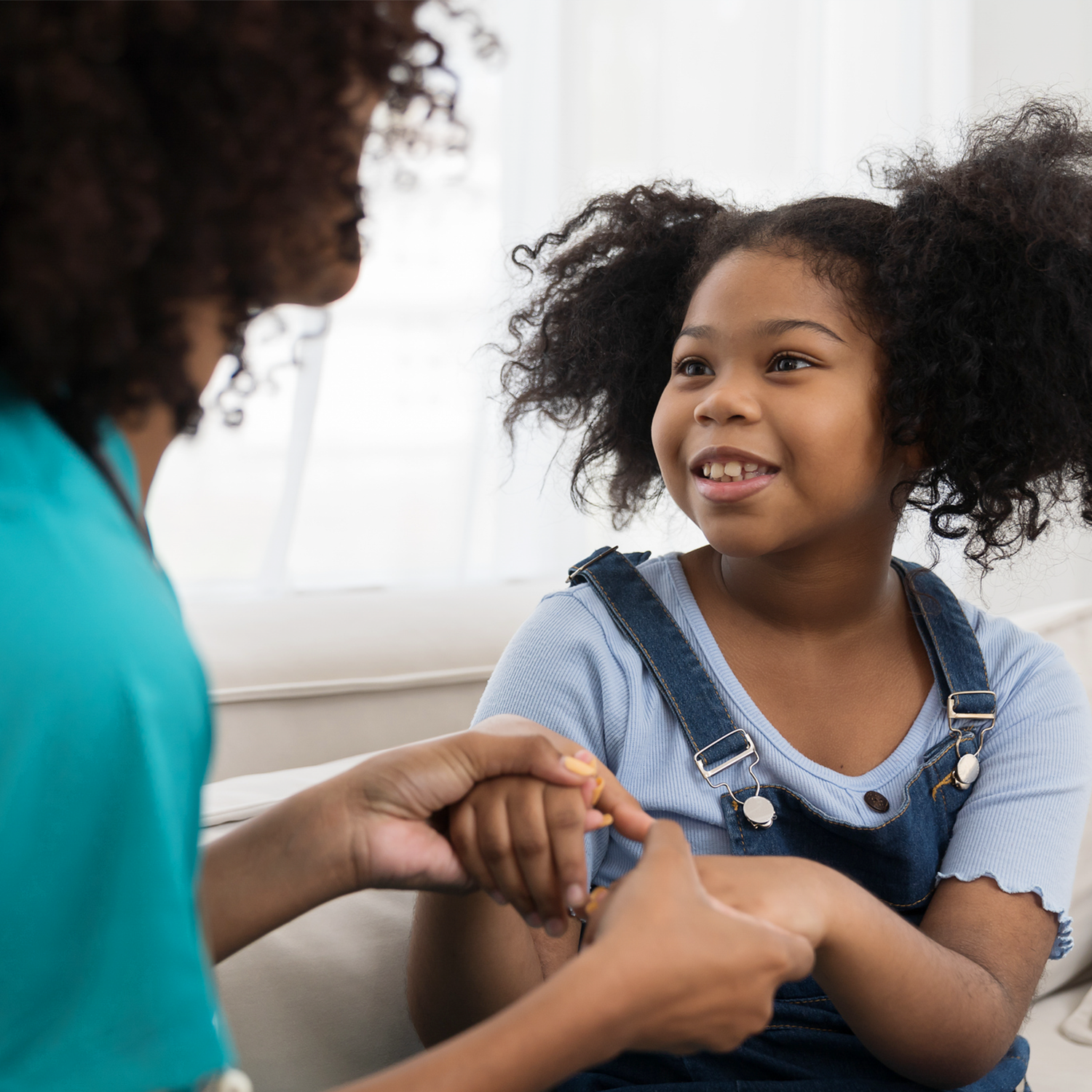 A young girl with curly hair smiling as she holds hands with an adult in a teal shirt, sitting inside near a window.