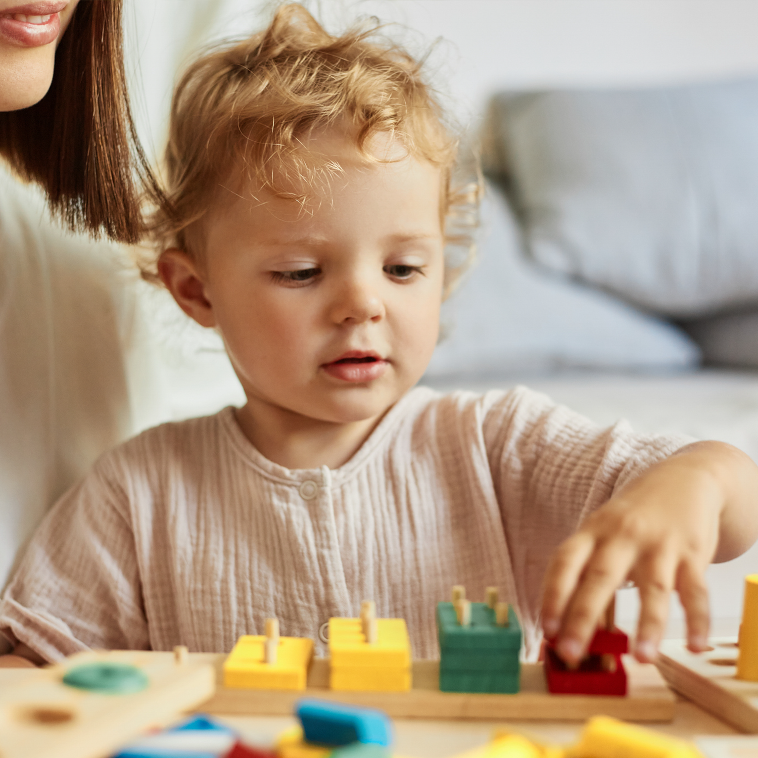 A young child playing with colorful building blocks at a table, with part of a woman's face visible in the background.