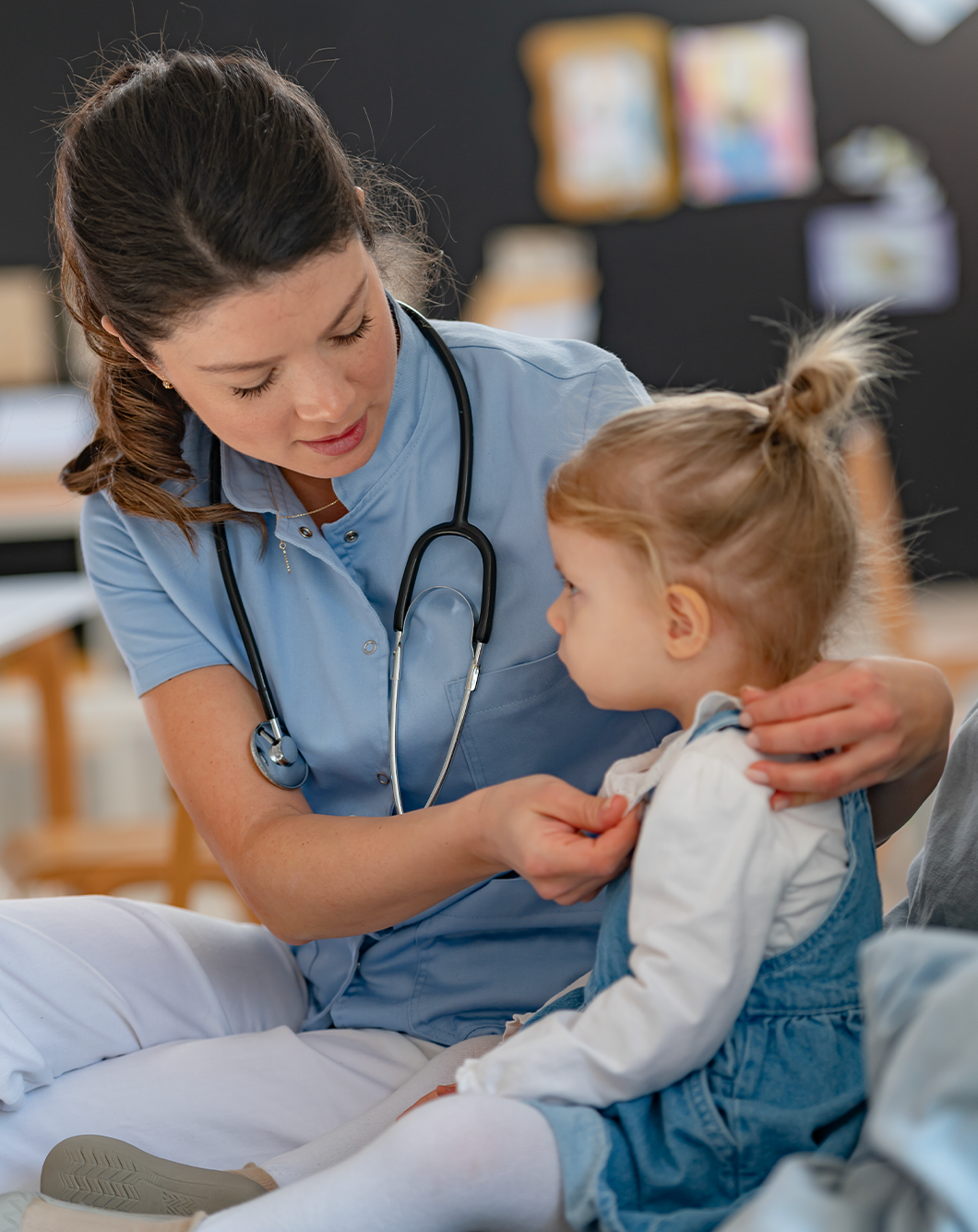 A female nurse in scrubs with a stethoscope around her neck gently examining a young girl in a hospital bed.