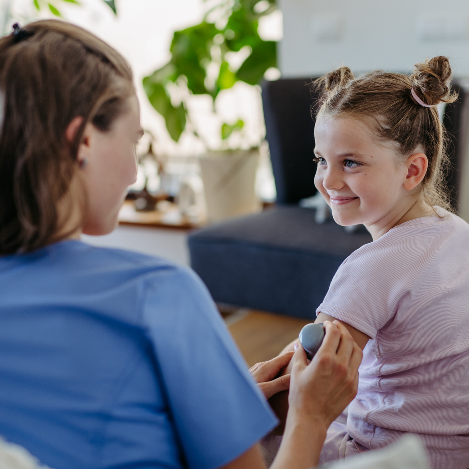 A young girl with light brown curly hair styled in two buns is smiling and sitting at a table during a check-up with a healthcare worker who is holding her arm with a blood pressure cuff. The healthcare worker is wearing a blue uniform, and the room has a large green plant in the background.