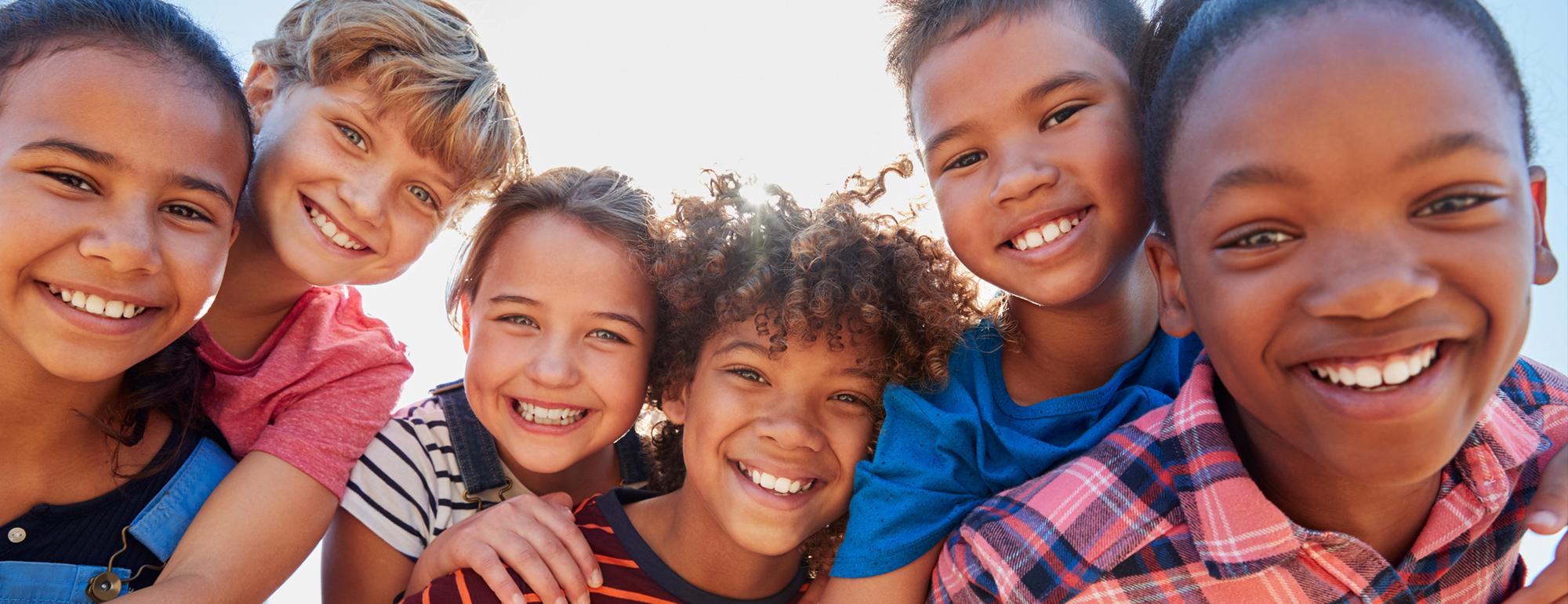 Group of seven kids smiling and looking at the camera outdoors, with sunlight shining behind them.