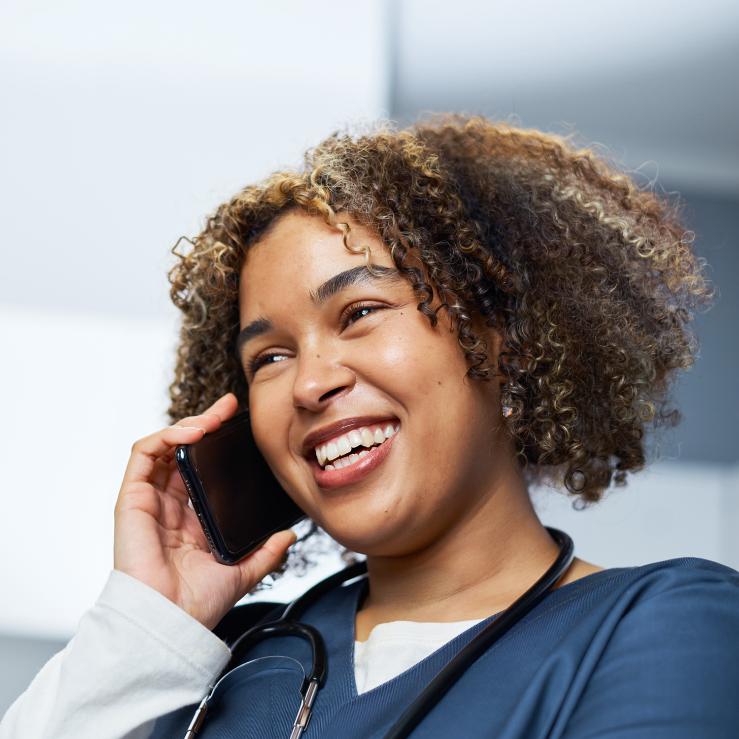 A smiling female healthcare worker in a navy scrub top talking on a mobile phone, with a stethoscope around her neck.