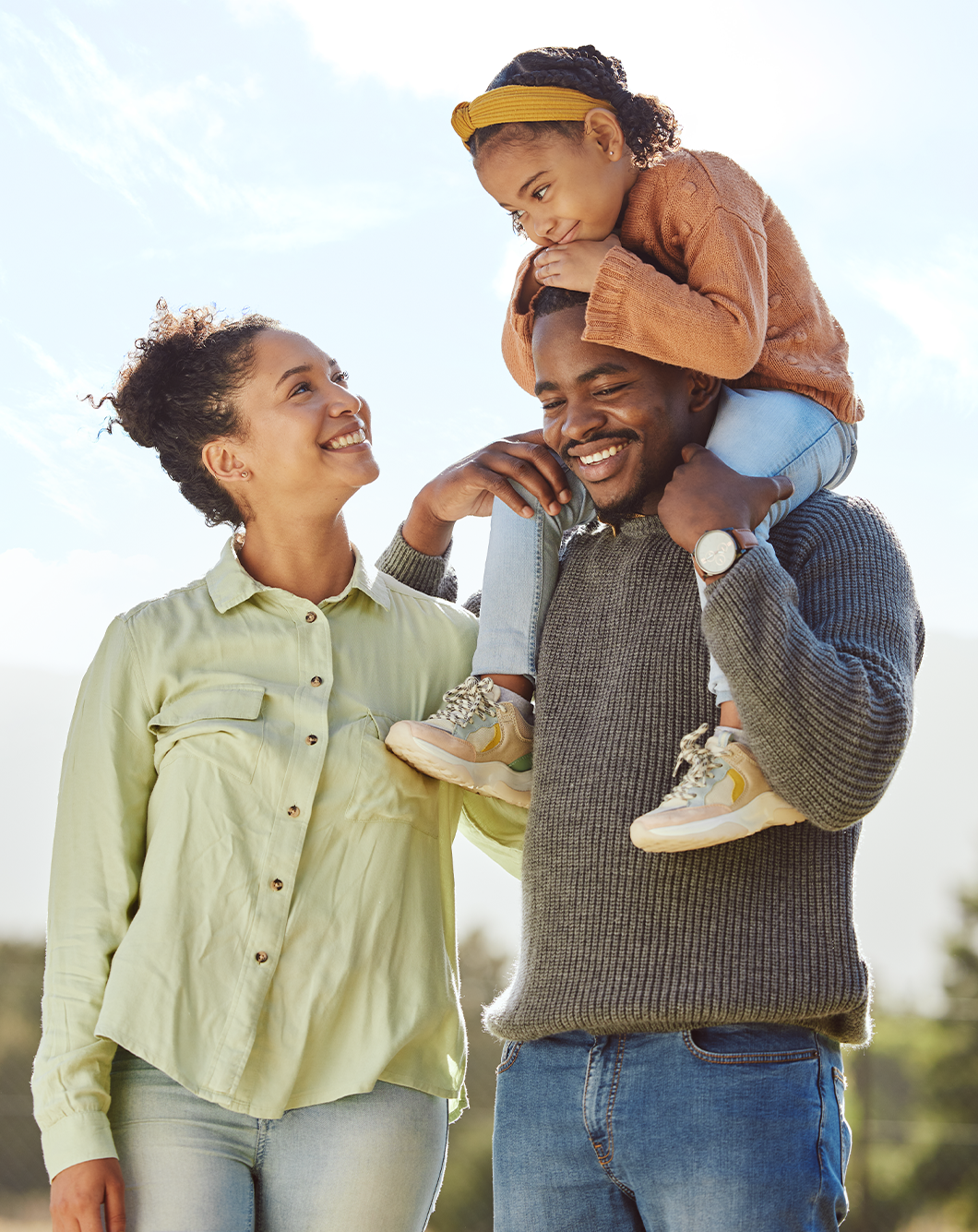 Family enjoying outdoor time, a woman and a man with a young girl on his shoulders, all smiling at each other.