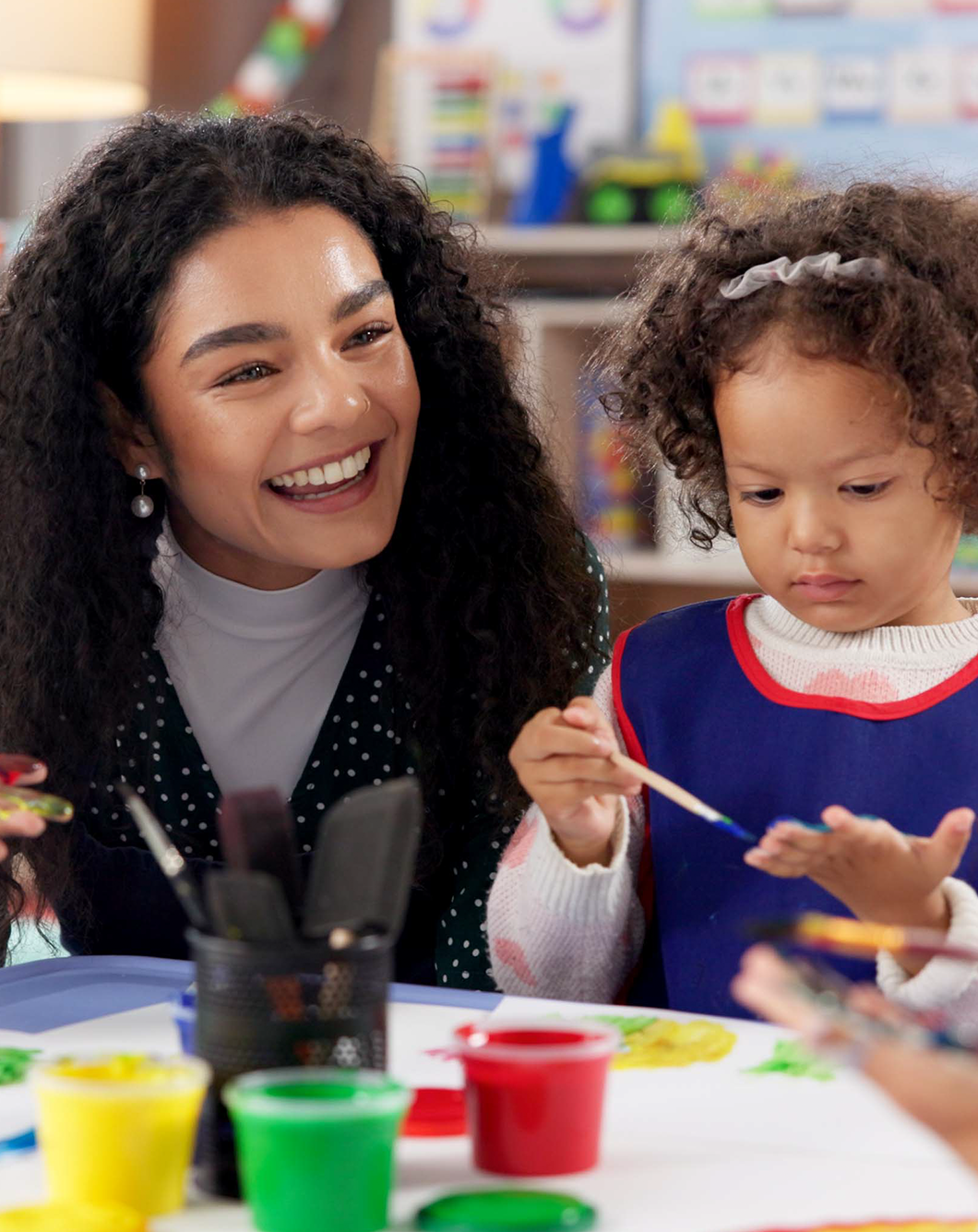 A woman with dark curly hair smiling while sitting at a table with a young girl painting with colorful paint in a classroom setting.