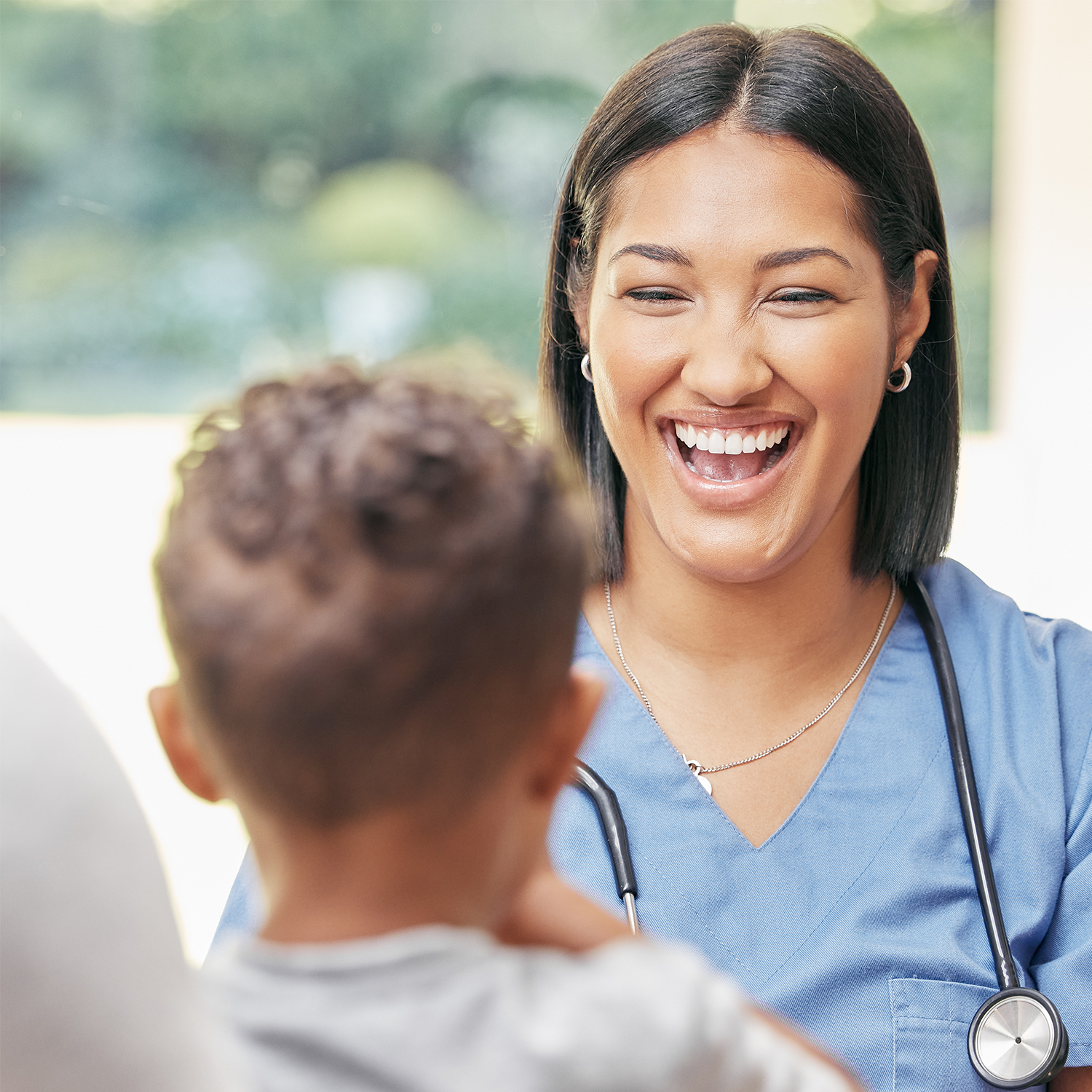 A female healthcare professional in blue scrubs smiling and talking to a young patient with curly hair.