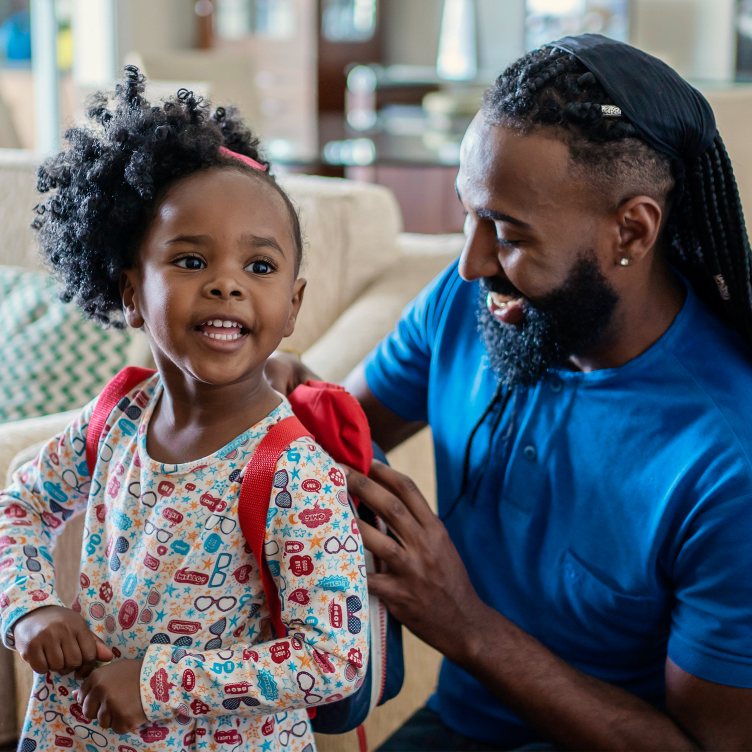 A young girl with a backpack talking to a man with a beard and braided hair, in a bright indoor setting.