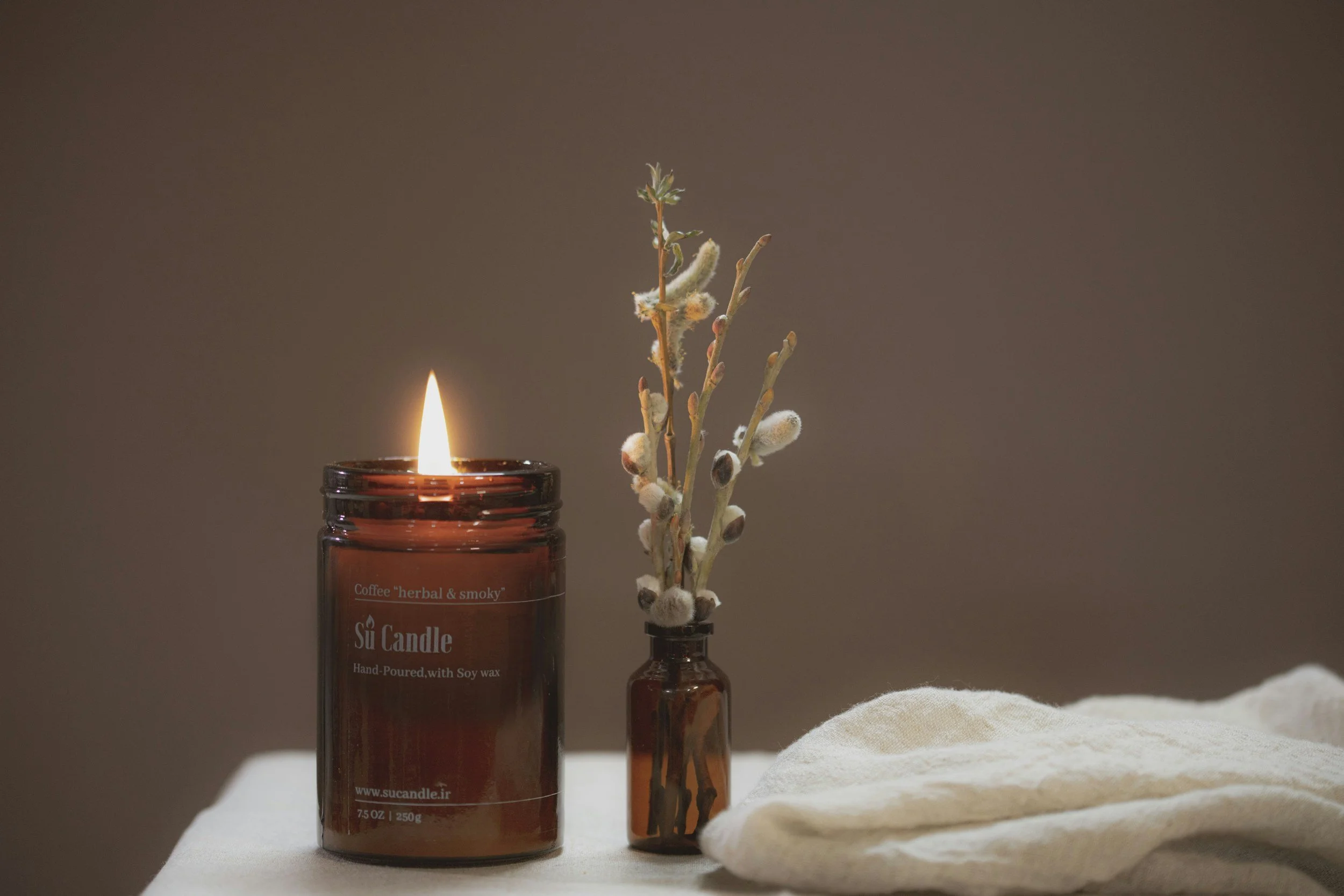 A lit brown glass candle jar with a label that reads 'Coffee herbal & smoky', placed beside a small brown glass vase containing pussy willow branches and a green sprig, with a white cloth and beige surface in the background.