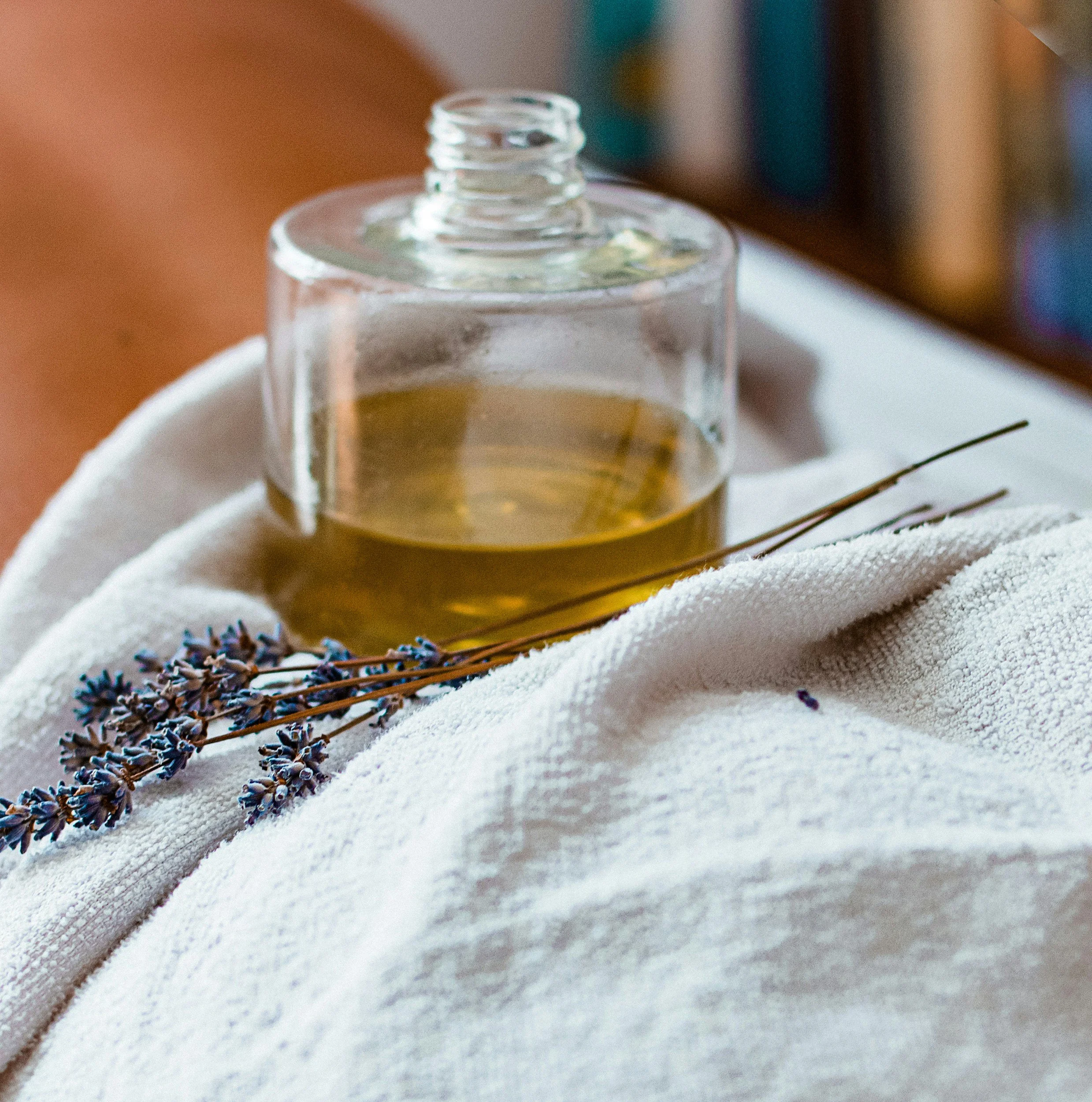 Close-up of a glass bottle with a narrow opening, placed on a white textured cloth with sprigs of lavender nearby, blurry background.