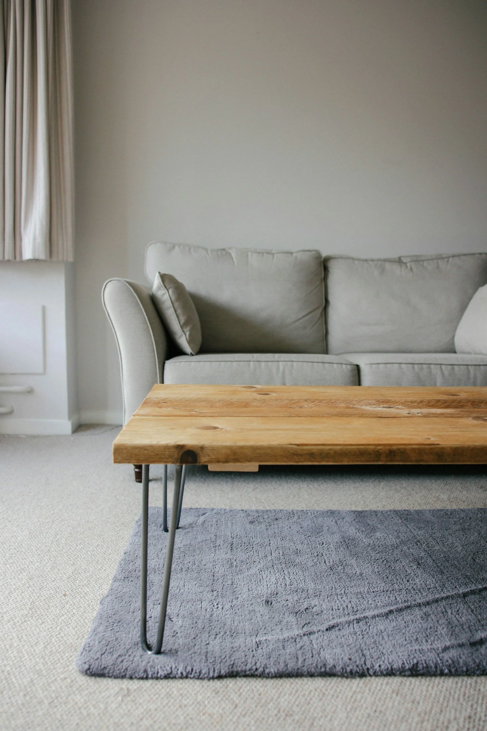 Living room with a beige sofa, a wooden coffee table, a gray rug, cream carpet, a wall with a window covered by beige curtains.