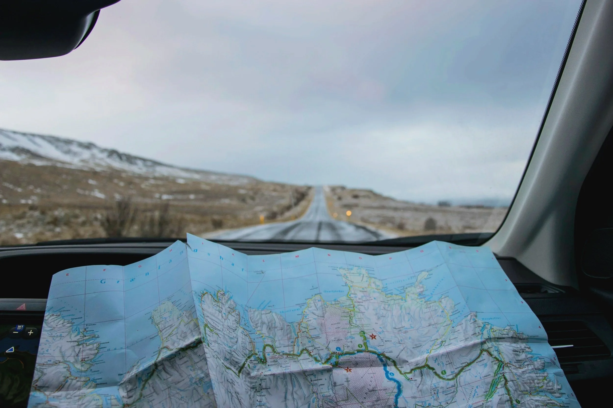 Inside a car, a map spread out on the dashboard with a scenic landscape of mountains and cloudy sky visible through the windshield.