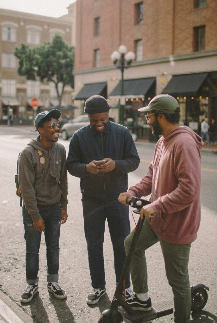 Three young men standing on a city sidewalk, smiling and laughing, with one of them holding a phone, two wearing hoodies and caps, and one riding an electric scooter, in front of a building with storefronts and a streetlamp.