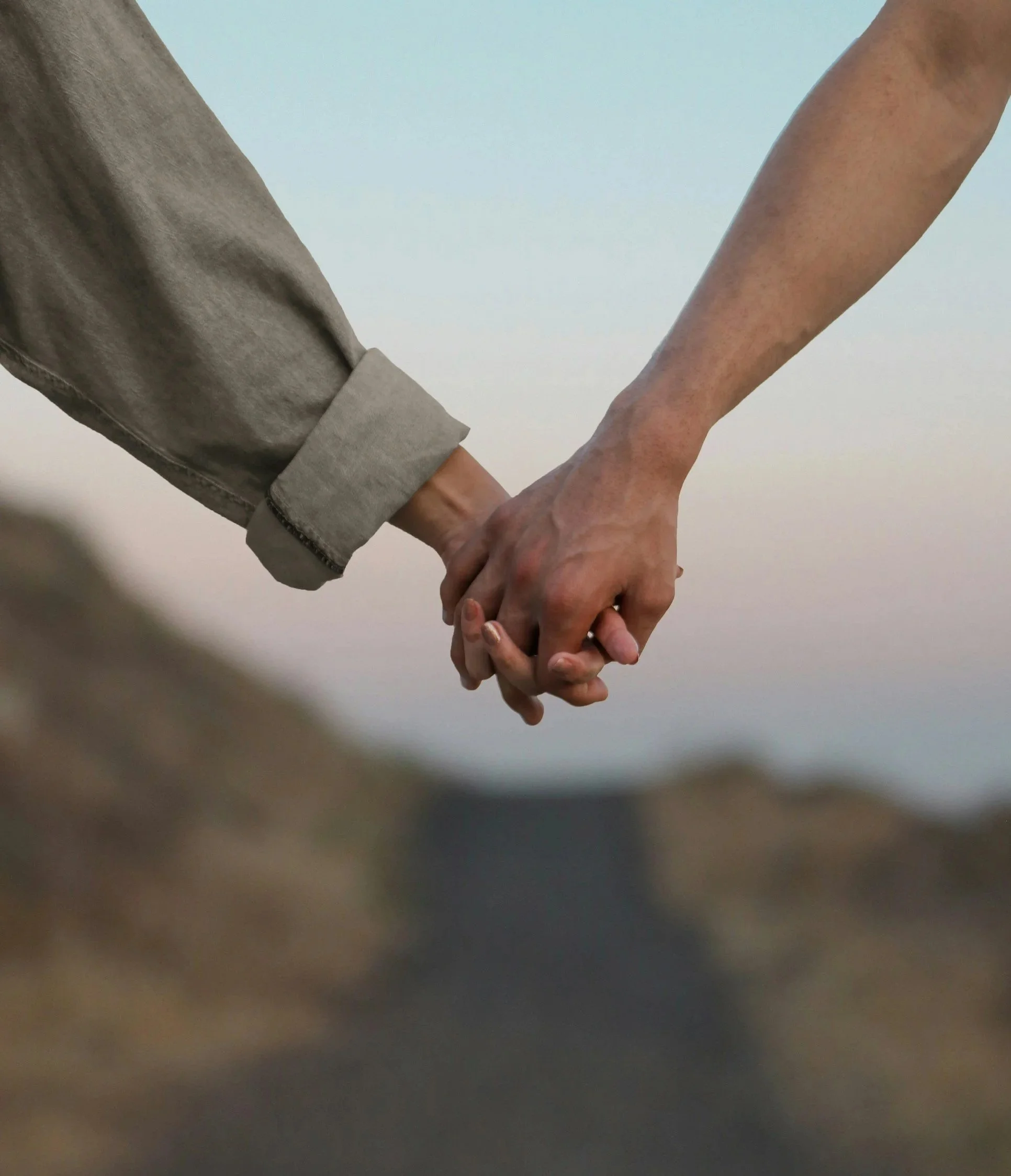 Two people holding hands, one person wearing a gray sleeve, the other with a tanned arm, against a light background.