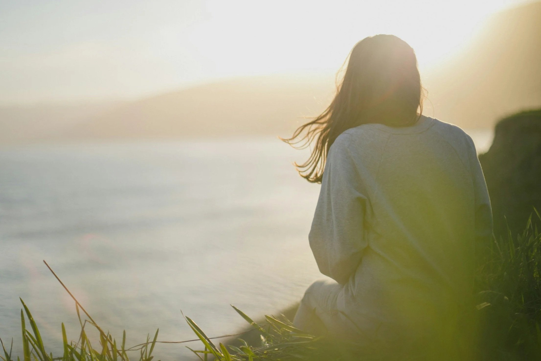 A person standing by water during sunset, with long hair partially covering their face, dressed in a light-colored sweatshirt and pants, surrounded by greenery.