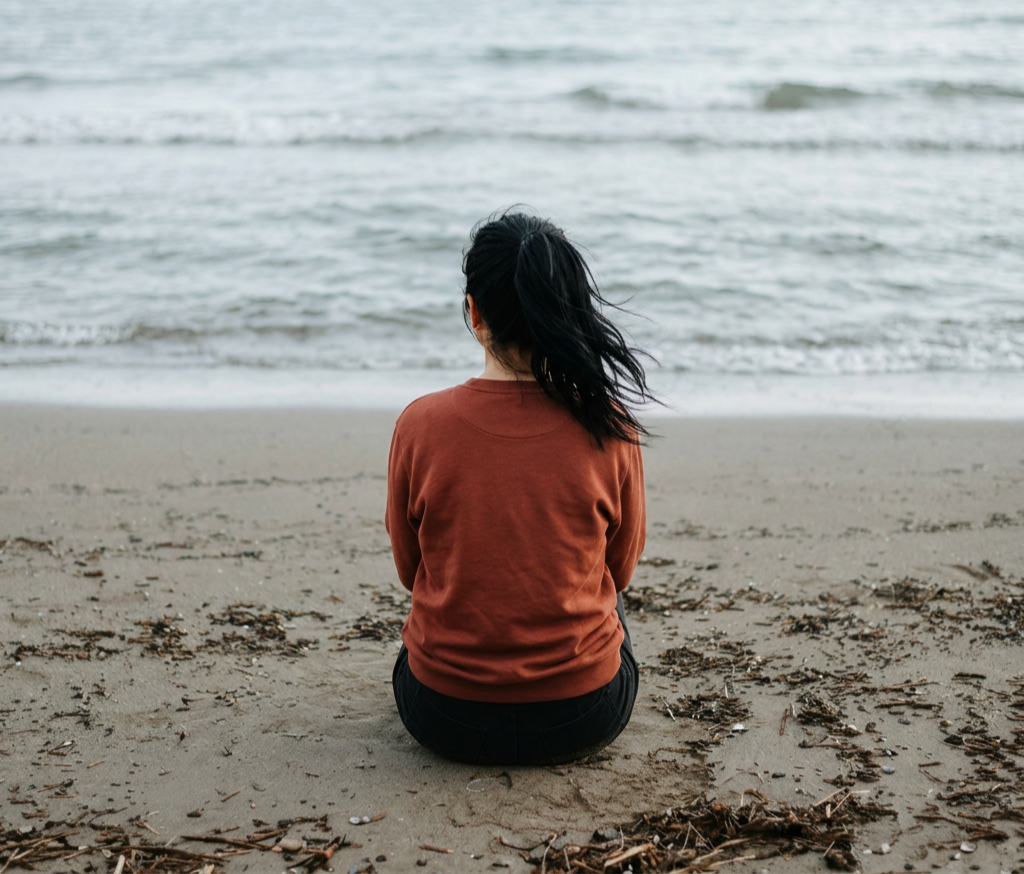 A woman with dark hair sitting on a sandy beach facing the water, wearing a brown sweatshirt.