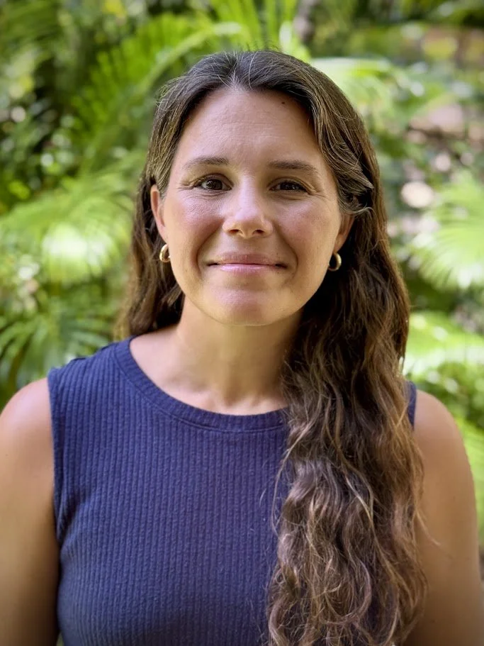 Portrait of a woman with long wavy brown hair, wearing a sleeveless blue top, standing outdoors with lush green foliage in the background.