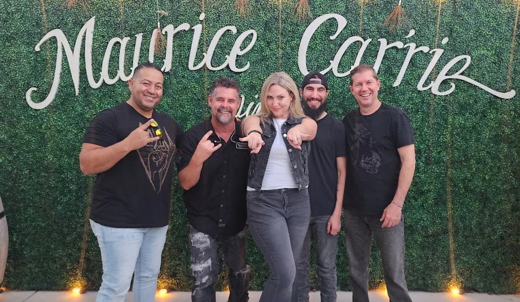 Group of five people standing in front of a green leafy backdrop with 'Maurice Corrib' written in cursive, posing and smiling for the camera.
