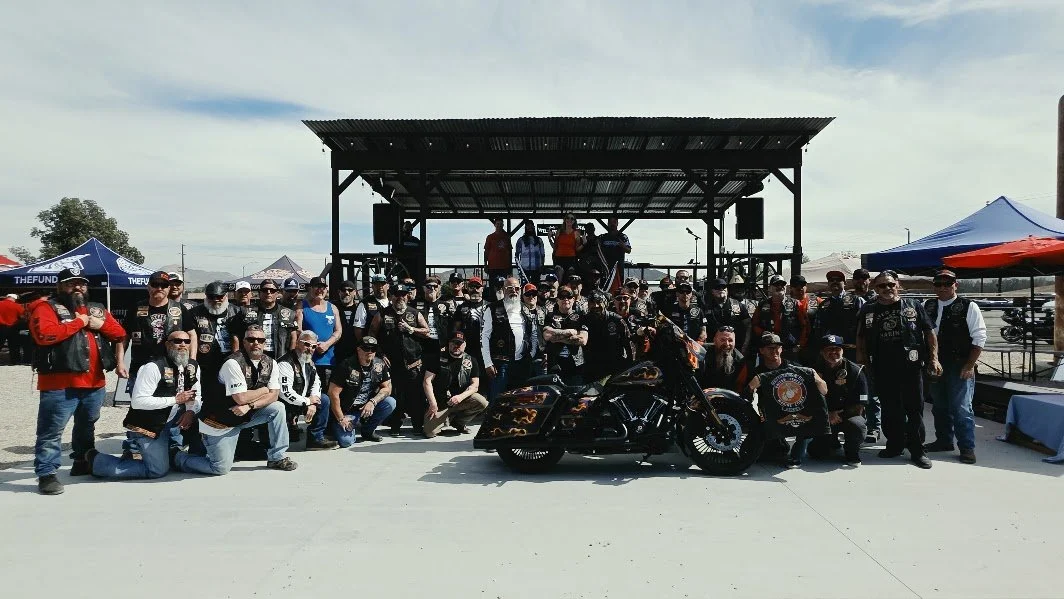 Group of motorcycle riders and enthusiasts gathered in front of a stage with a custom black motorcycle with flames painted on it, during an outdoor event.