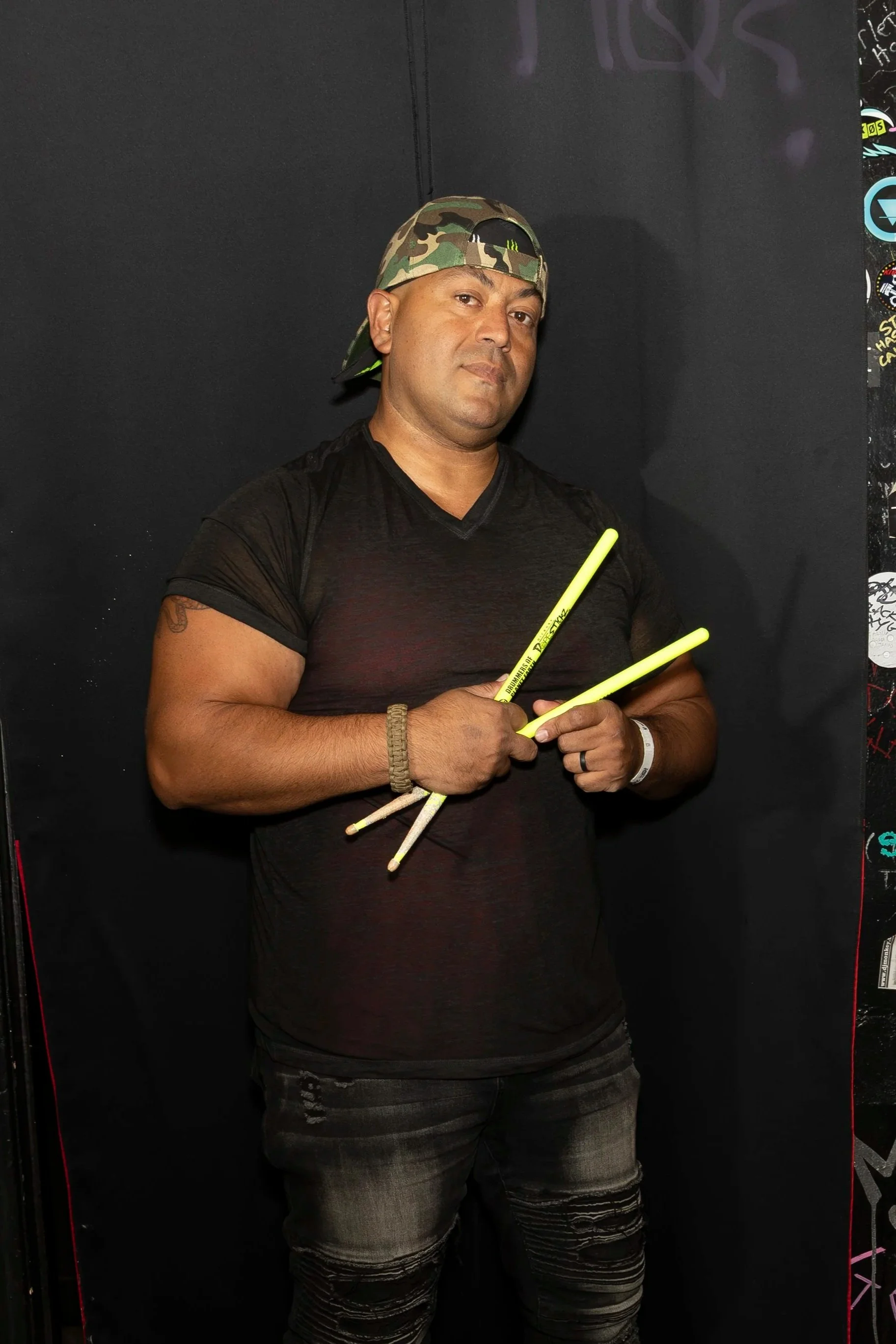 A man with tan skin, wearing a camo baseball cap backward and a black t-shirt, is holding glowing yellow drumsticks. He is standing against a black background with some graffiti and stickers visible on the right side.