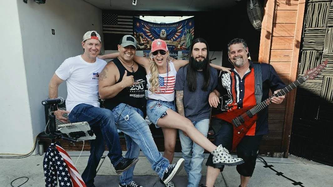 Five people standing together, smiling, with musical instruments and patriotic accessories, in front of a backdrop with an eagle and American flags.