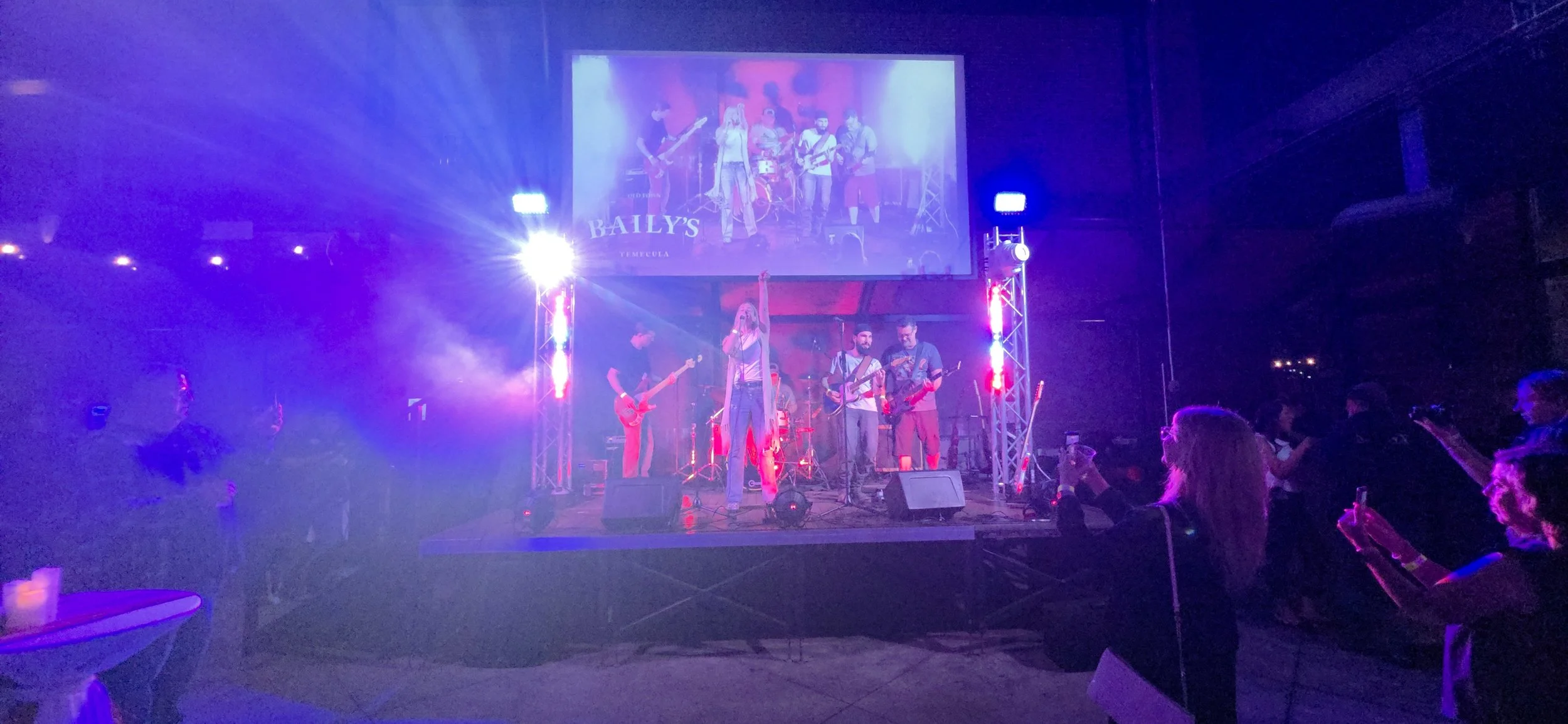 A live band performing on stage at a concert with vibrant purple and pink lighting, with people in the audience watching and taking photos.