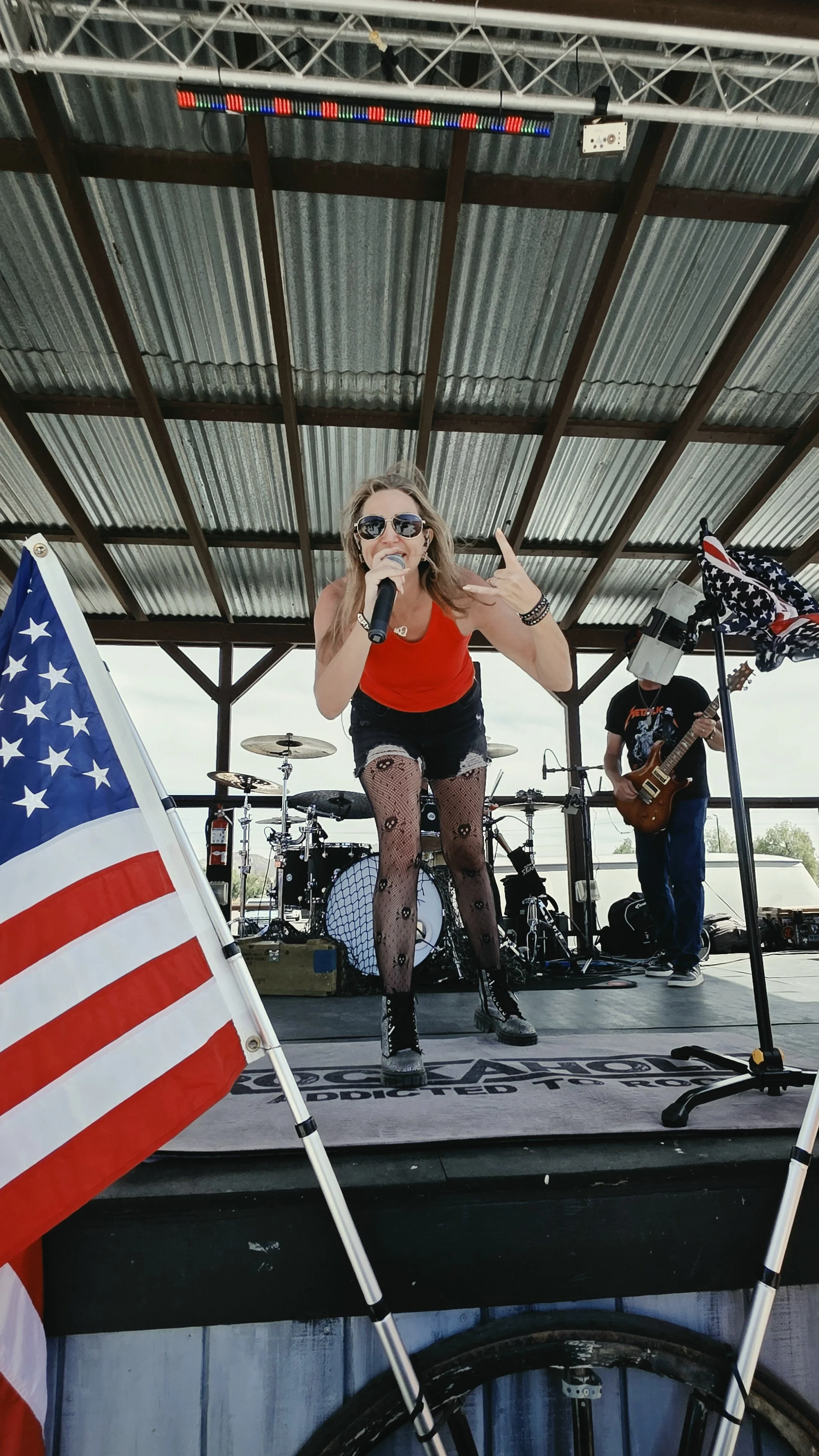 A woman singing into a microphone on stage, wearing sunglasses, a red top, fishnet stockings with skull patterns, and black combat boots, with gloves and bracelets. A guitar player is in the background, and American flags are displayed on the stage.