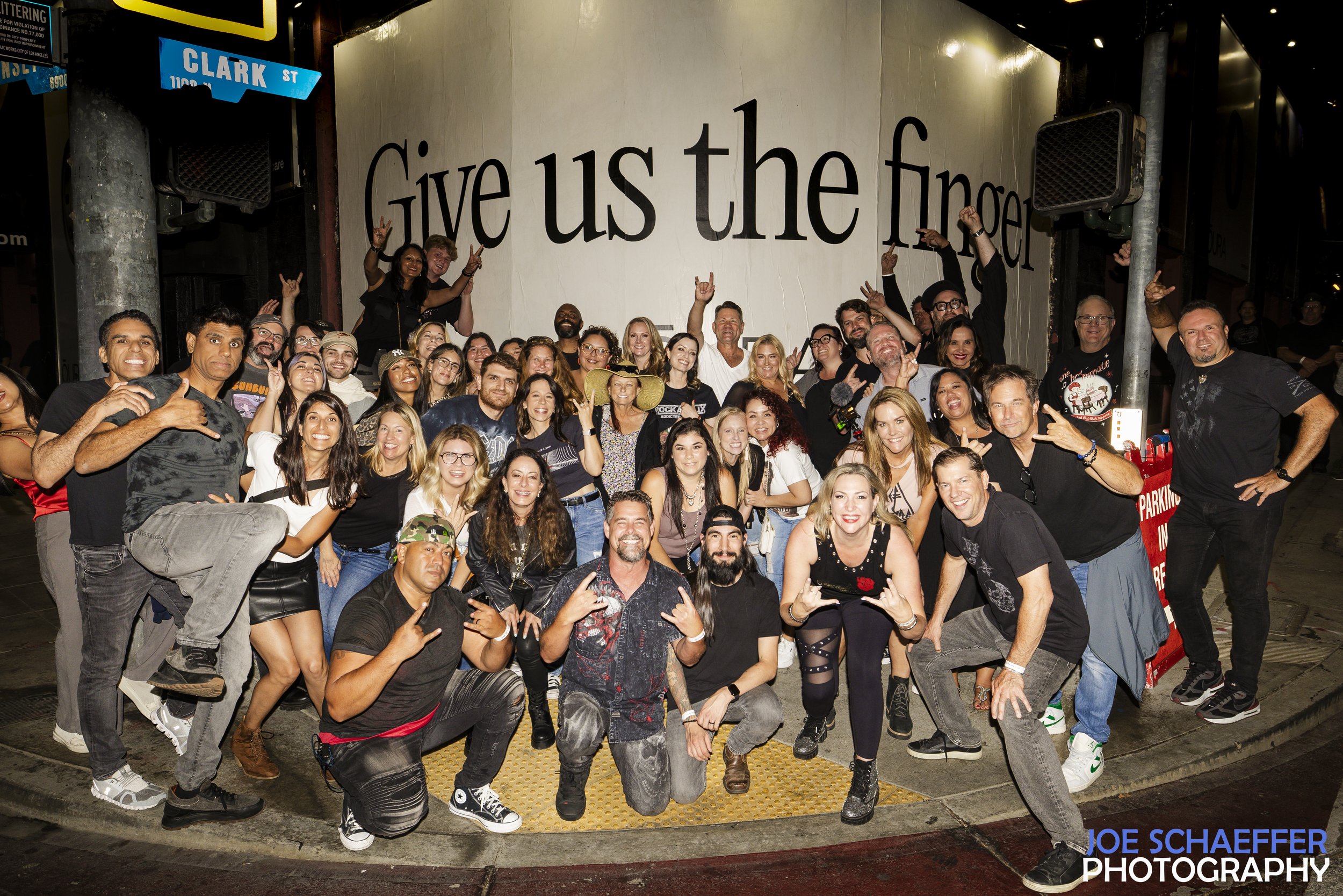 A large group of people, smiling and making gestures, standing outside at night under a sign that says 'Give us the finger.' The group appears cheerful and diverse, with some individuals making rock and peace signs. The setting looks like urban street or alley.