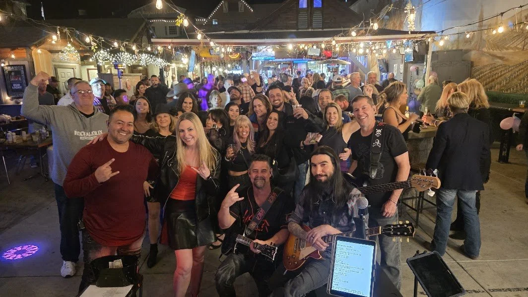 Group of people at a bar or music venue, some holding guitars and others smiling or making gestures, with string lights overhead and a crowded background.