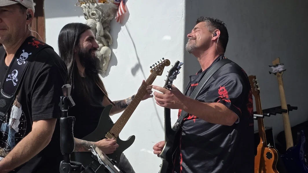 Three men in a music studio or garage, two playing guitars and one partially visible on the left, all appearing to enjoy playing music together.