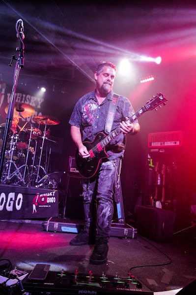 A man playing an electric guitar on stage during a live music performance with colorful stage lighting.