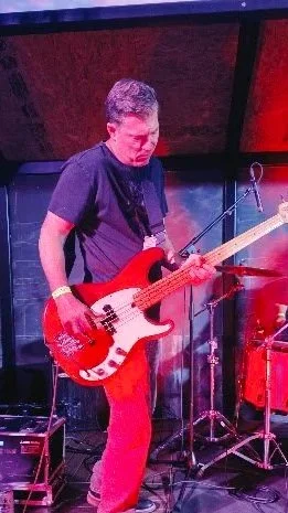 A person playing a red electric bass guitar on stage, with musical equipment and a drum set in the background.