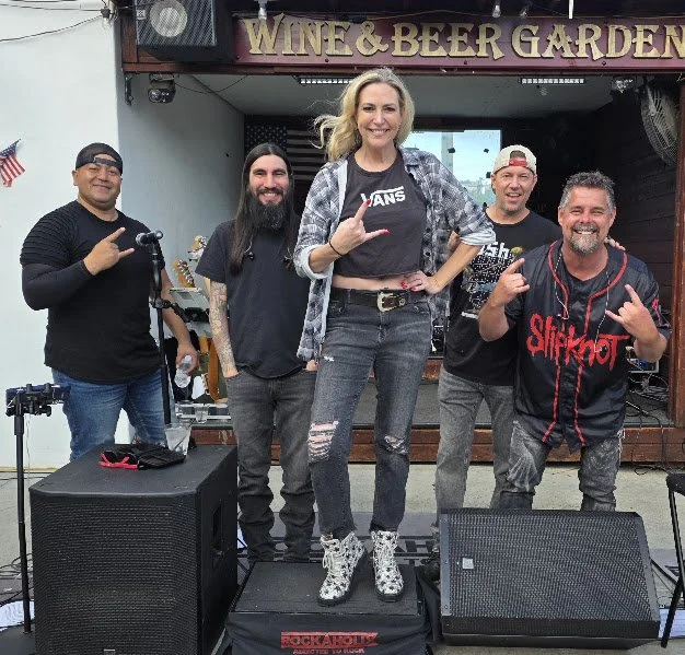 Group of five people standing on a stage in front of a sign that reads 'WINE & BEER GARDEN,' with musical equipment around them. They are smiling, making rock hand signs, and dressed casually.
