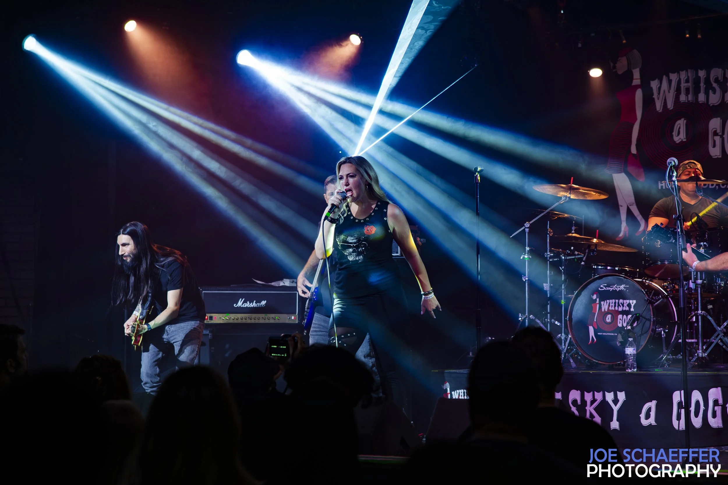 A live band performs on stage with colorful lighting, including a female singer at the center holding a microphone, a guitarist to her left, and a drummer in the background. The backdrop features the words 'Whisky a Go Go' and a silhouette of a person. Audience members are visible at the bottom of the image.