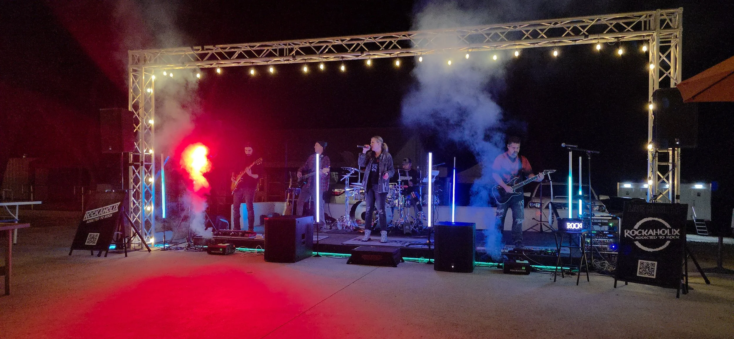 A band performing on an outdoor stage at night with colorful lights, smoke effects, and musical instruments.