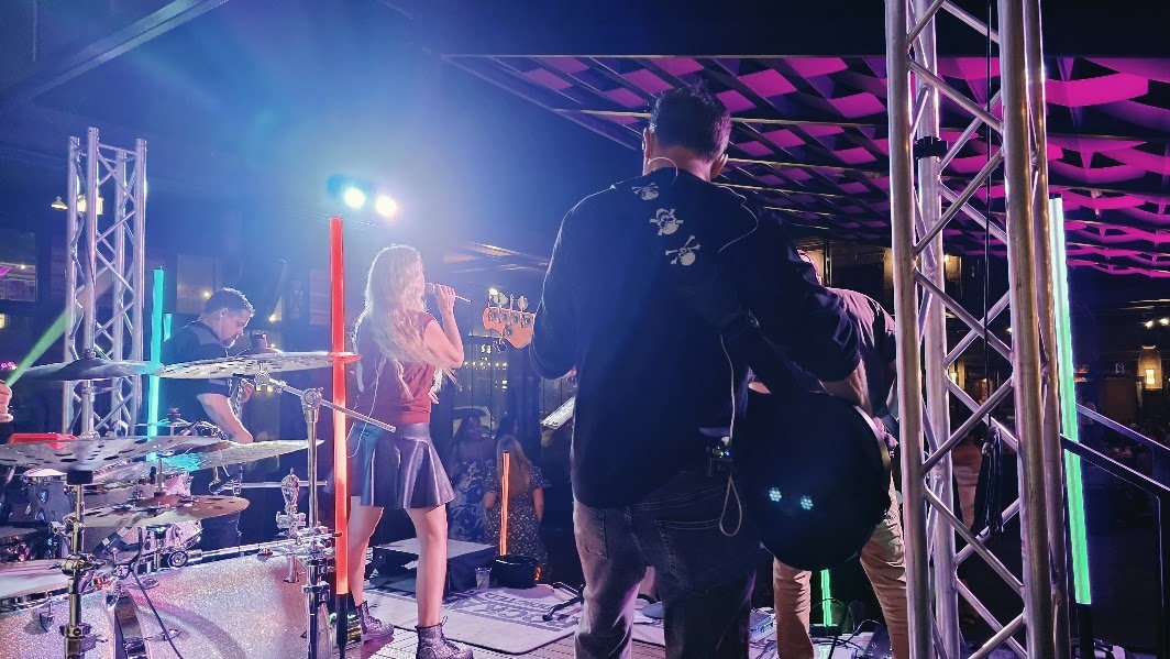 A live music performance on a stage with colorful lighting, featuring a female singer, musicians playing instruments, and an audience in the background.