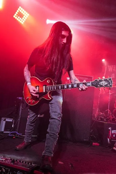 Musician with long hair playing an electric guitar on stage with red lighting.