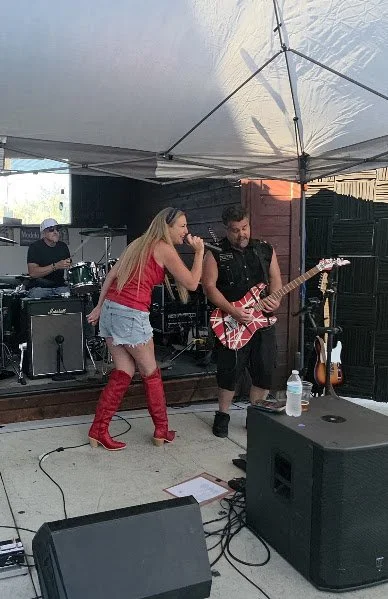 A band performing outdoors under a canopy, including a female singer in red top and denim shorts with red boots, a male guitarist in black shirt, and a drummer in the background.
