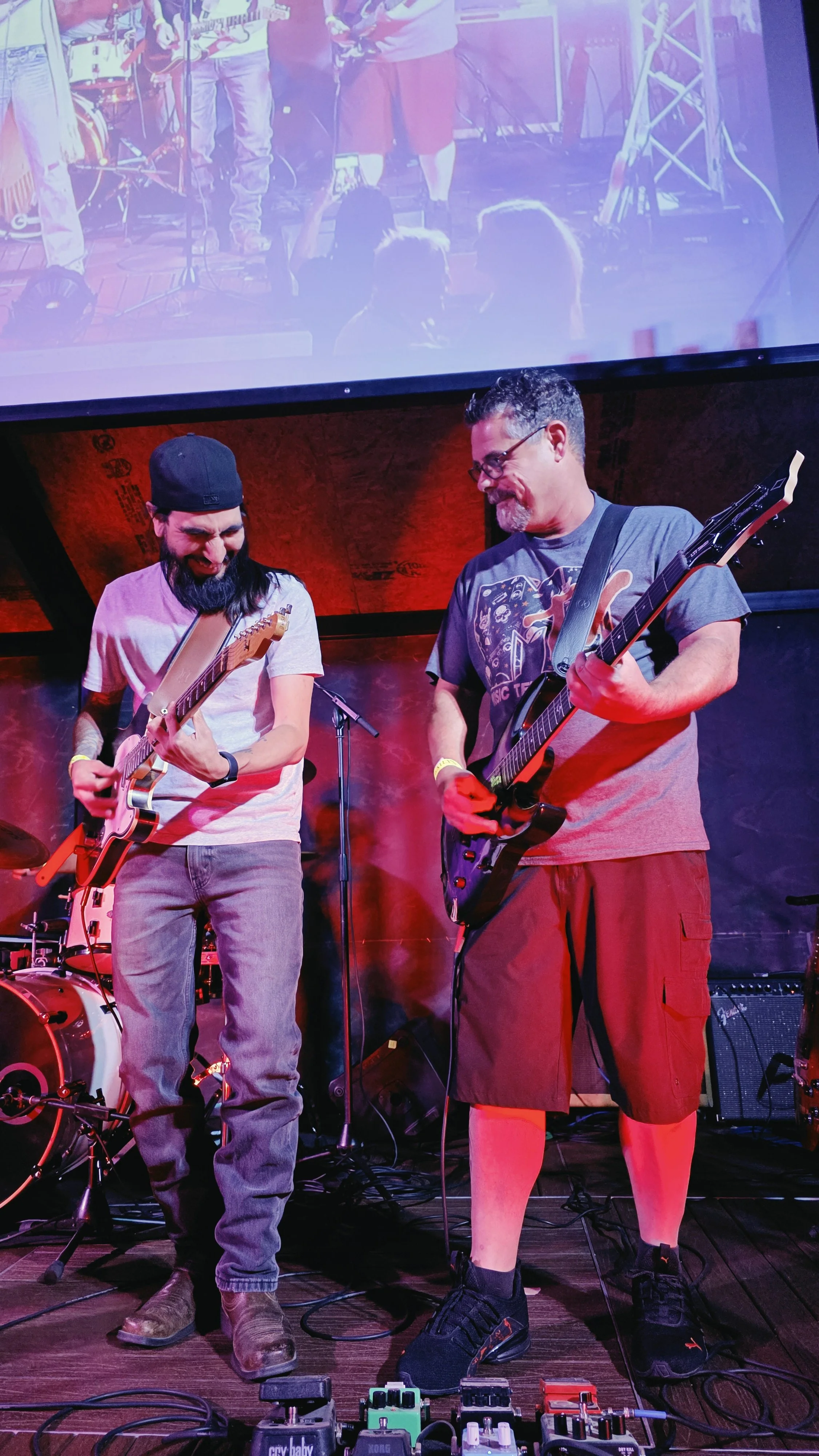 Two men playing electric guitars on a stage with musical equipment and a drum set in the background. One man is wearing a white T-shirt, jeans, and a backward cap; the other is wearing a gray T-shirt, red shorts, and black sneakers. A large screen ab
