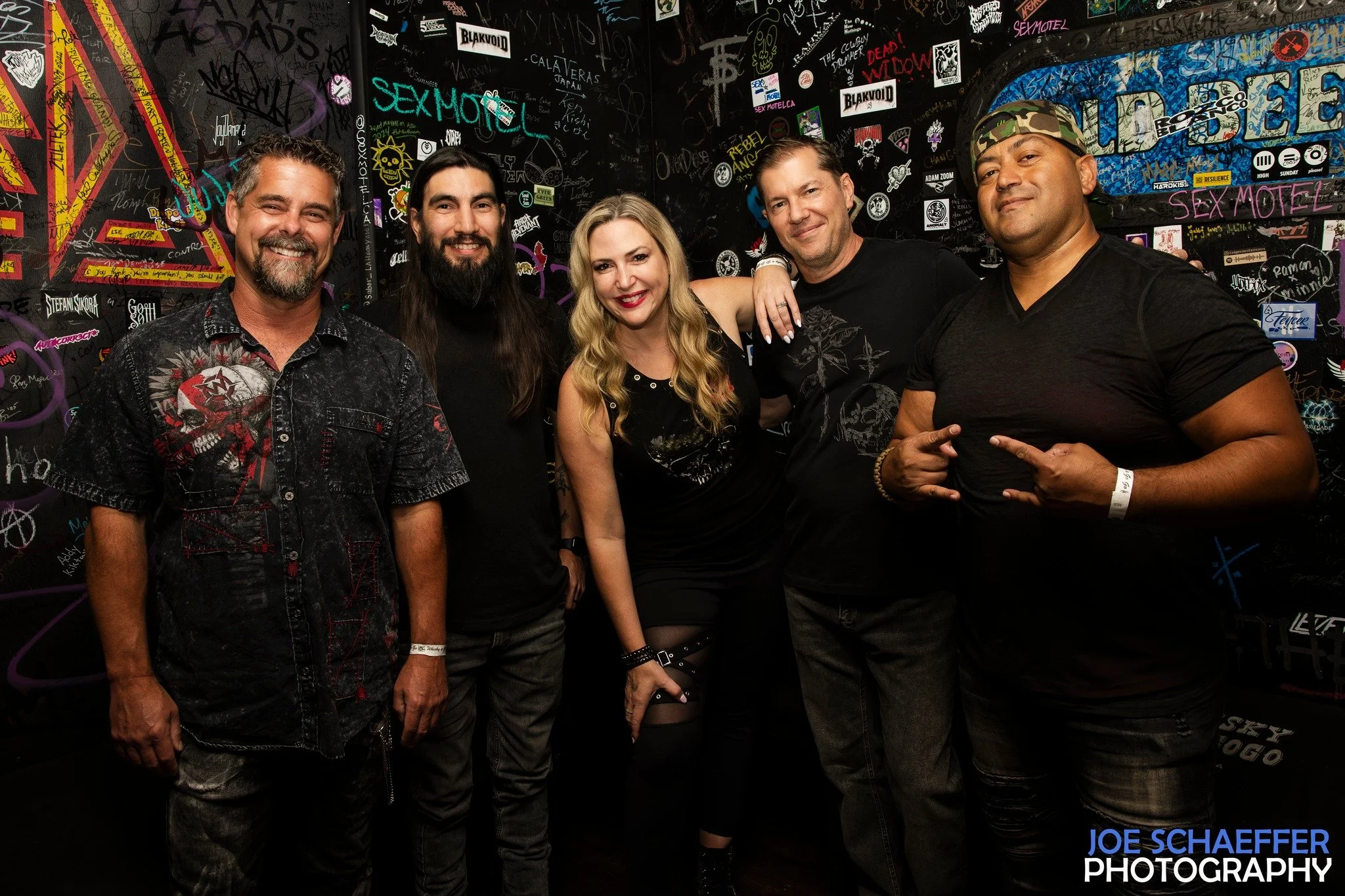 Group of five people posing in front of a graffiti-covered wall at a nightclub or music event, with stickers and graffiti tags in the background.