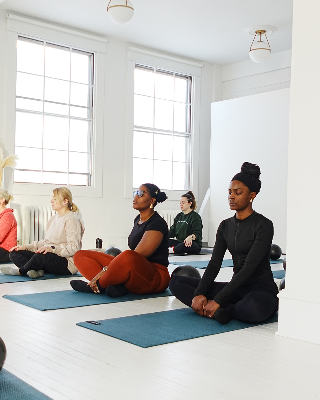 A group of women practicing meditation or yoga in a bright, clean room with large windows and white walls, sitting cross-legged on mats with their eyes closed or relaxed.