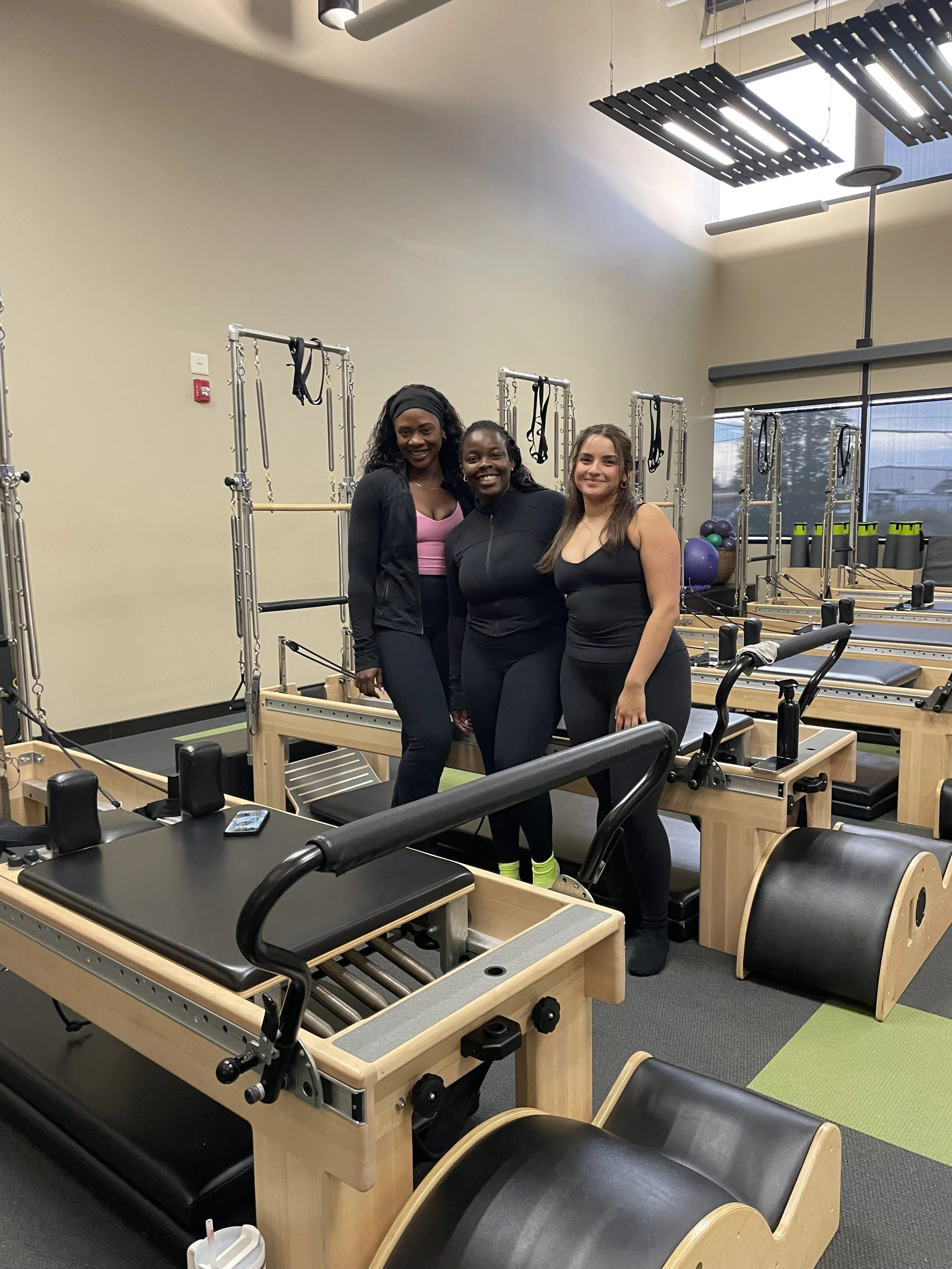 Three women stand in a Pilates or fitness studio with reformer machines, colorful exercise balls, and water bottles on the equipment. They are smiling and posing for a photo.