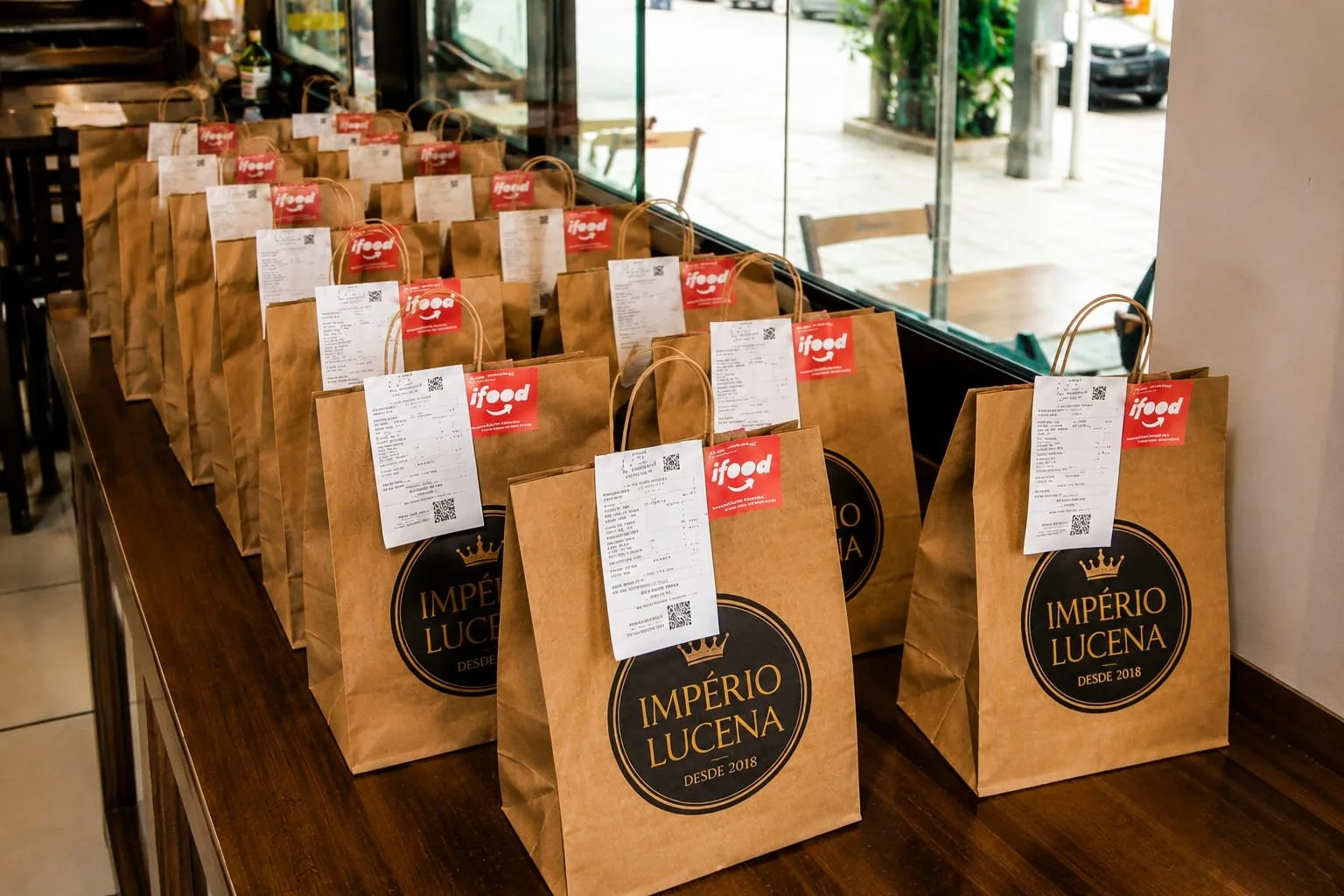 Sacolas de papel com o logo Império Lucena e print das comandas da comida em uma mesa de restaurante.