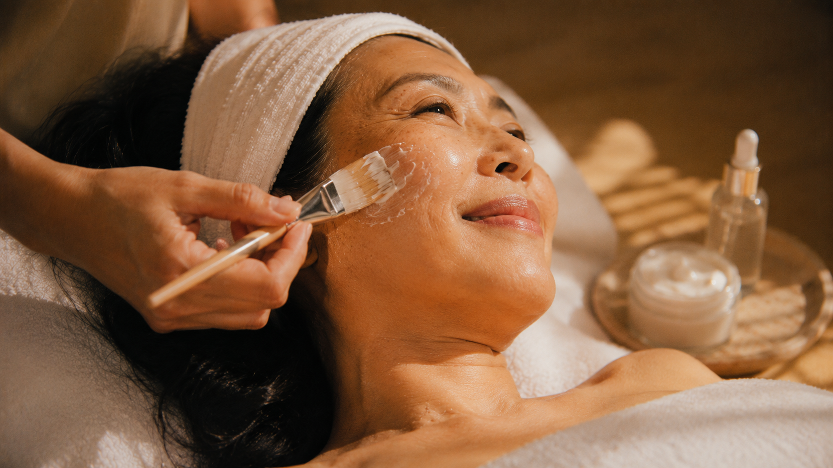 A woman lying on a spa bed during a facial treatment, wearing a white headband, with a skincare product being applied to her face using a brush, and a tray with skincare bottles and jars nearby.
