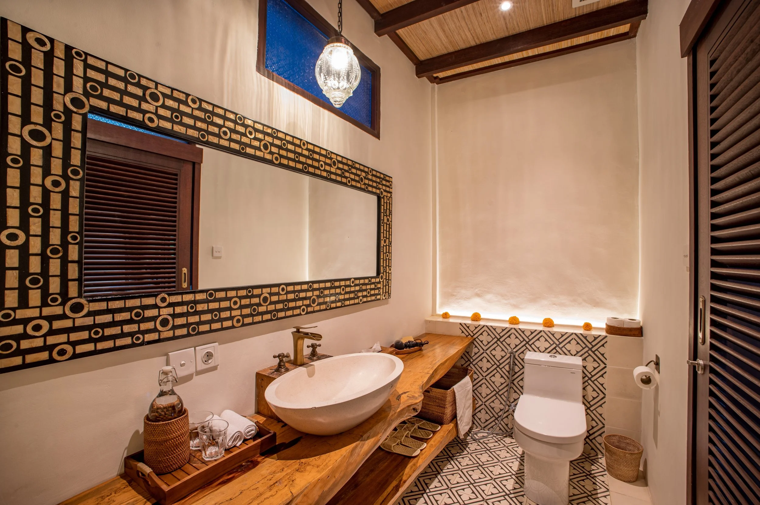 A bathroom with a large mirror framed in black and gold geometric design, a vessel sink with a wooden countertop, a decorative chandelier hanging from a wooden beamed ceiling, a small window with blue glass, and patterned grey and white floor tiles. 