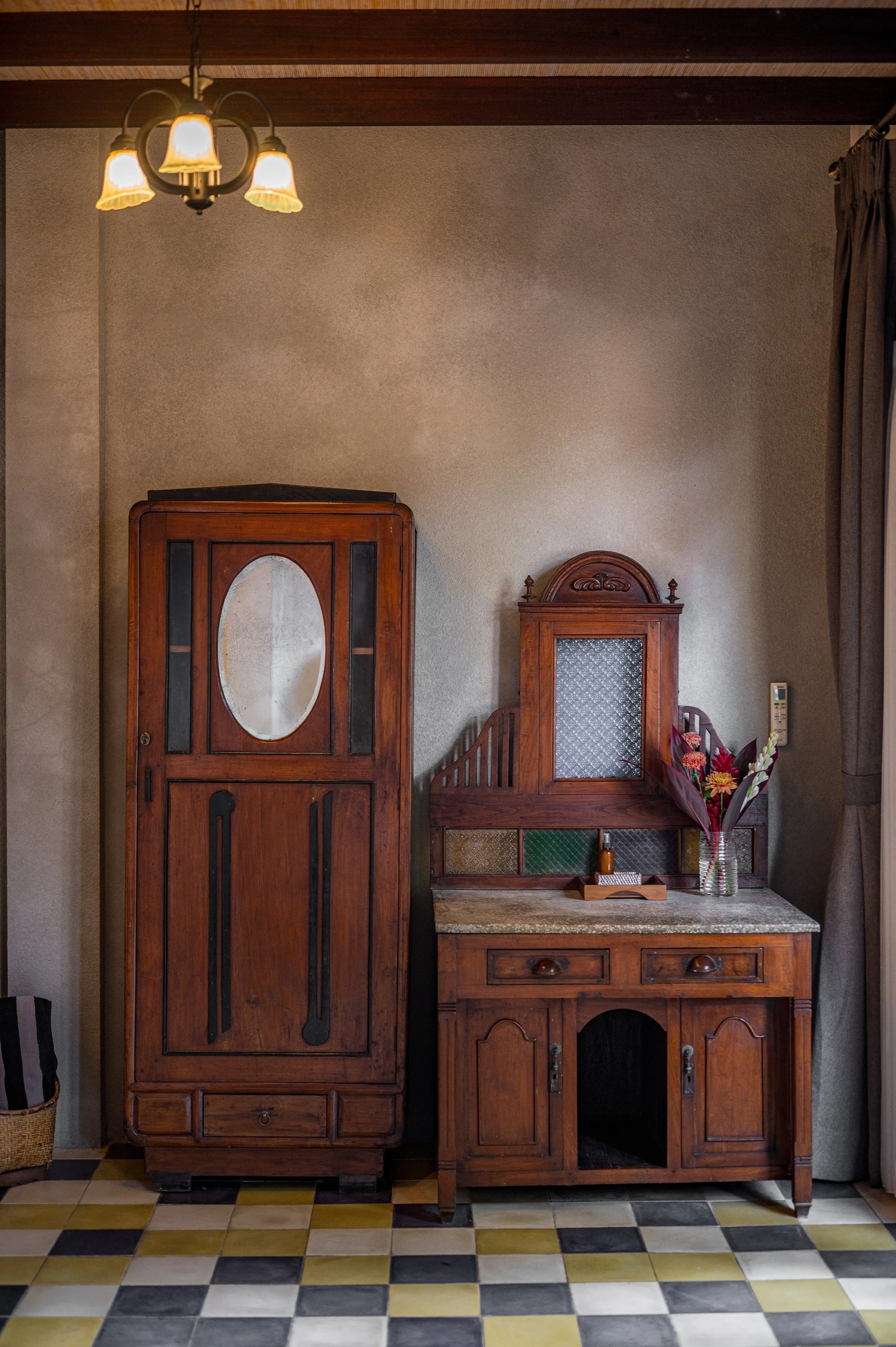 Room with wooden furniture including two cabinets and a countertop, a vase with flowers, colorful tiled floor, beige textured wall, ceiling with dark wood beams, and a ceiling lamp with four lampshades.